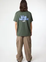 Camiseta de manga corta color verde militar, de corte regular fit y confeccionada en algodón orgánico. Presenta un estampado gráfico grande en la espalda con el texto 'blue banana' en tonos celestes y oscuros, acompañado de un pequeño recuadro blanco con inscripciones.