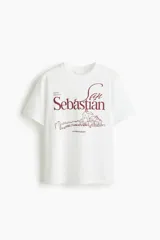 Remera de algodón blanca de corte holgado, con cuello redondo, mangas cortas y estampa frontal en color bordó con la inscripción "San Sebastián" y un dibujo lineal de un paisaje costero.
