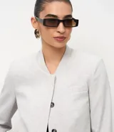 Chaqueta de sastre estructurada color gris claro, con cuello redondo tipo rectilínea, cierre frontal con botones y mangas largas con botones en los puños. Presenta un corte levemente corto y diseño minimalista.