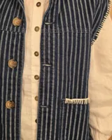 Chaleco de denim color marrón, confeccionado en 100% algodón, con bolsillos frontales amplios, cierre de botones y costuras decorativas bordadas en blanco en los bordes y los bolsillos.