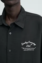 Camisa de corte relajado confeccionada en tejido fluido con mezcla de viscosa, color negro. Presenta cuello solapa, manga corta, cierre frontal con botones y un bordado a contraste en el pecho.