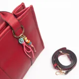 Cartera tipo tote de color rojo con textura granulada, doble asa superior y correa larga ajustable extraíble. Incluye un colgante decorativo con cuentas coloridas y tiras de cuero sintético en uno de los laterales.