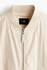 Campera bomber en mezcla de algodón y nylon color beige, con cuello elevado acanalado y cierre frontal de cremallera. Cuenta con bolsillos laterales y un bolsillo con cierre en la manga izquierda. Puños y bajo en punto acanalado, de corte holgado.