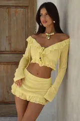 Conjunto de dos piezas color amarillo, compuesto por un top de manga larga con escote off-shoulder, volados y cierre frontal con lazos, y una minifalda a juego con detalle de volado en el ruedo.