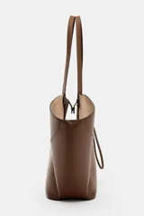 Bolso de hombro tipo maxi shopper color marrón, con doble asa y estructura flexible que se pliega hacia afuera. Presenta un detalle de tira ajustable con cordón en el centro frontal. Incluye un compartimento interior extraíble con cierre de cremallera y cierre principal mediante botón imantado.
