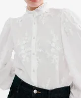 Blusa blanca de algodón con bordados florales, cuello alto con volados y mangas largas abullonadas con puños ajustados.