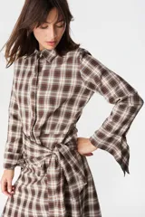 Camisa de corte holgado con estampado de cuadros en tonos marrones y beige. Presenta cuello clásico, cierre frontal con botones y mangas largas con aberturas laterales que permiten anudar la prenda en la cintura.