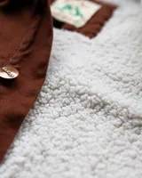 Campera estilo workwear de Denali, confeccionada en canvas resistente color marrón. Presenta interior forrado en corderito sherpa, cuello de pana negro, cierre frontal con botones metálicos color cobre y cuatro bolsillos frontales con costuras reforzadas en contraste.