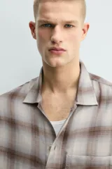 Camisa relaxed fit de manga larga con cuello solapa, confeccionada en tejido con mezcla de viscosa. Presenta un bolsillo plastrón en el pecho, bajo con aberturas laterales y cierre frontal de botonadura. El diseño es a cuadros en tonos beige y marrón.