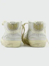 Championes de caña media Golden Goose modelo Mid Star, color blanco con efecto desgastado. Presentan una estrella lateral con glitter plateado, detalles de perforaciones tipo brogue y suela con acabado envejecido.