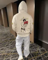 Canguro con capucha color beige, de corte regular, con estampado estilo pixel art en la espalda que representa a un boxeador con guantes rojos y estrellas alrededor. Presenta detalles de estrellas estampadas en las mangas.