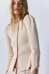 Chaqueta de punto acanalado color beige, con cuello redondo, cierre frontal de botones y mangas largas con hombros abullonados y terminación asimétrica.