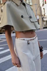 Trench corto tipo capa de algodón en color beige, con cuello solapa, trabillas en los hombros, bajo redondeado y cierre frontal cruzado con botones.