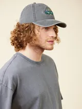 Gorro de gabardina gris oscuro con visera curva, ajustable en la parte trasera con hebilla metálica. Presenta bordado frontal con el logo de la marca y el año 1985.