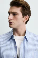 Camisa de corte regular fit confeccionada en tejido de algodón, con diseño de rayas verticales en tonos azules. Presenta cuello abotonado, manga larga con puños abotonados, cierre frontal de botones y un bolsillo en el pecho.