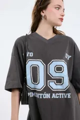 Remera oversize gris oscuro con cuello en V y mangas cortas. Presenta una estampa frontal estilo universitario con el número "09" y las inscripciones "MVD", "UY" y "PIMENTÓN ACTIVE" en tonos celestes y blancos.