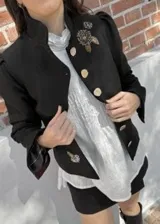 Chaqueta de paño de lana y cashmere color verde oscuro, de corte estructurado con cuello mao, mangas largas con hombros abullonados y puños con tajo. Cuenta con cierre frontal de botones dorados grandes y detalles de bordados dorados artesanales en el pecho y el bajo.