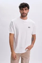 Remera blanca de manga corta con cuello redondo y una pequeña etiqueta estampada en el pecho con diseño de montaña y texto.