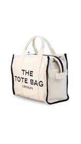 Bolso tipo tote bag de lona color beige con detalles de ribetes negros. Presenta el texto 'THE TOTE BAG' y 'LINCOLN'S' estampado en el frente. Cuenta con doble asa superior y correa ajustable extraíble.