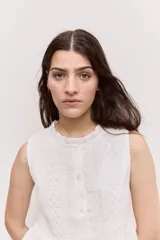 Blusa sin mangas de color blanco, con cuello redondo fruncido y cierre frontal de botones. Combina un cuerpo texturizado con una parte inferior de encaje calado y diseño peplum.