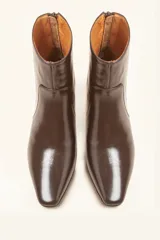 Bota de caña corta confeccionada en cuero vacuno color chocolate con acabado brillante. Presenta punta fina, cierre posterior y taco chupete bajo.