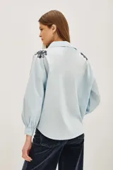 Camisa blanca de algodón con cuello camisero y mangas largas abullonadas con puños ajustados. Presenta detalles bordados en los hombros con motivos vegetales y aplicaciones brillantes.