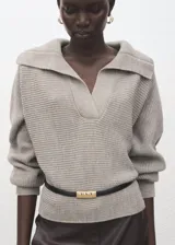 Jersey de punto color beige, con cuello polo, escote en V y mangas largas con puños ajustados. Presenta una textura acanalada y un diseño holgado.