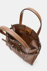 Bolso tipo mini shopper de cuero sintético color marrón caramelo, con costuras blancas visibles. Posee asas de mano y una correa bandolera ancha de tela color beige. El frente tiene un detalle de tira con hebilla metálica dorada.