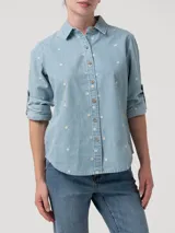 Camisa de manga larga de denim celeste claro, con bordados pequeños en forma de copo de nieve o flor blanca distribuidos por toda la prenda. Posee cuello camisero, cierre frontal con botones marrones y mangas que se pueden enrollar y sujetar con una tira.