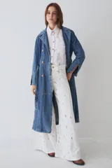 Trench largo de denim azul, con diseño de doble botonadura, solapas clásicas y mangas largas con puños ajustables. Presenta un corte recto y estructurado.