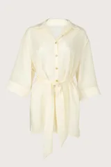Camisa larga fluida de color crema con cierre frontal de botones, mangas tres cuartos anchas y cinturón ajustable para anudar en la cintura.