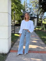 Blusa blanca de algodón con textura arrugada, cuello redondo, mangas largas abullonadas con puños elásticos y cierre frontal con botones y volantes.