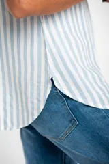 Camisa oversize celeste y blanca a rayas verticales, con cuello clásico, mangas cortas con dobladillo y cierre frontal con botones.
