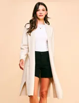 Cardigan largo de tejido color beige claro, sin cuello, con bolsillos frontales y detalles de pespuntes decorativos a lo largo del borde frontal.