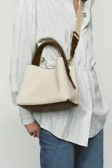 Bolso tipo saca color crudo con cuerpo en tejido canvas, asa corta de mano trenzada en marrón y correa bandolera adicional. Cuenta con cierre de botón imantado y bolsillo interior con cremallera.