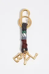 Llavero con mosquetón y anilla metálica dorada, cuerpo trenzado con secciones de cordón negro, tejido estampado a rayas y cuero marrón. Incluye tres dijes metálicos dorados con las letras "B", "Y" y "L".