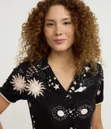 Conjunto de pijama de dos piezas confeccionado en viscolycra, compuesto por una blusa de manga corta con cuello solapa y botones frontales, y un short con cintura elástica y lazo. Estampado integral de temática espacial con estrellas, planetas y explosiones en tonos rosados y blancos sobre fondo negro.