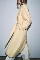 Abrigo largo color crema confeccionado en mezcla de lana. Diseño oversize con cuello en V, mangas largas y cinturón ajustable con lazada.