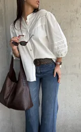 Camisa blanca de estilo romántico con cuello mao, botones frontales y mangas tres cuartos abullonadas con detalle de frunces. Presenta un diseño con textura sutil y costuras decorativas en la parte superior delantera.