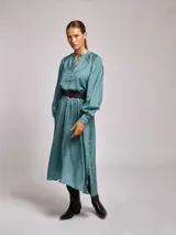 Vestido midi de satén con estampado geométrico en tonos marrones y naranjas. Presenta cuello mao, mangas largas abullonadas con puños ajustados y un cinturón a tono con hebilla metálica que marca la cintura.