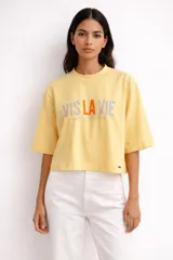 Remera de corte boxy y mangas cortas, color amarillo pastel. Presenta un estampado frontal con la frase 'VIS LA VIE' en letras grises y naranjas.