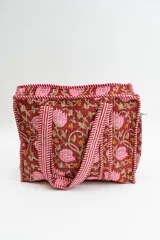Bolso tipo tote acolchado con estampado floral en tonos rojizos, rosados y verdes. Presenta asas de mano con diseño de rayas verticales en color rojo y blanco, y ribetes a juego en los bordes.