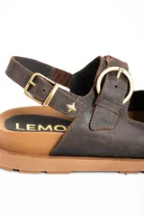 Sandalias de cuero marrón oscuro, con doble tira ancha en el empeine y tira en el talón, todas con hebillas metálicas doradas. Cuenta con suela plana y gruesa de tono tostado con estriado inferior.
