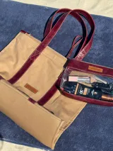 Bolso tipo tote de lona color beige con asas de cuero color bordó, cierre superior y bolsillo interno.