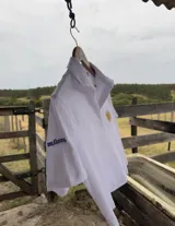 Remera tipo polo blanca de manga corta con cuello y abertura frontal. Presenta un bordado del Sol de Mayo en el pecho y la palabra "milong" bordada en azul en la manga izquierda.