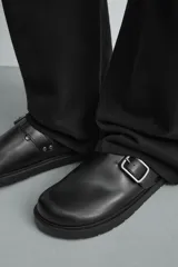 Zueco destalonado de cuero negro con suela de plataforma plana y detalle de hebilla metálica plateada en el empeine.
