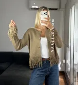 Cárdigan de tejido de punto color crema, con cuello redondo, mangas largas y cierre frontal con botones metálicos. Presenta detalles de flecos en los puños y en el ruedo.