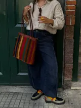 Cartera tipo tote de cuero con diseño de franjas verticales multicolores en tonos tierra, rojo y azul. Cuenta con correas trenzadas de cuero y cierre superior.