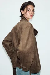 Chaqueta de gamuza sintética color marrón, con cuello subido, cierre frontal cruzado con botones y mangas largas anchas. Cuenta con bolsillos delanteros y cinturón ajustable del mismo tejido.
