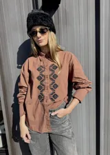 Camisa de algodón color marrón con bordado geométrico en color negro en la parte frontal. Presenta cuello clásico, cierre frontal con botones y mangas largas con puños.
